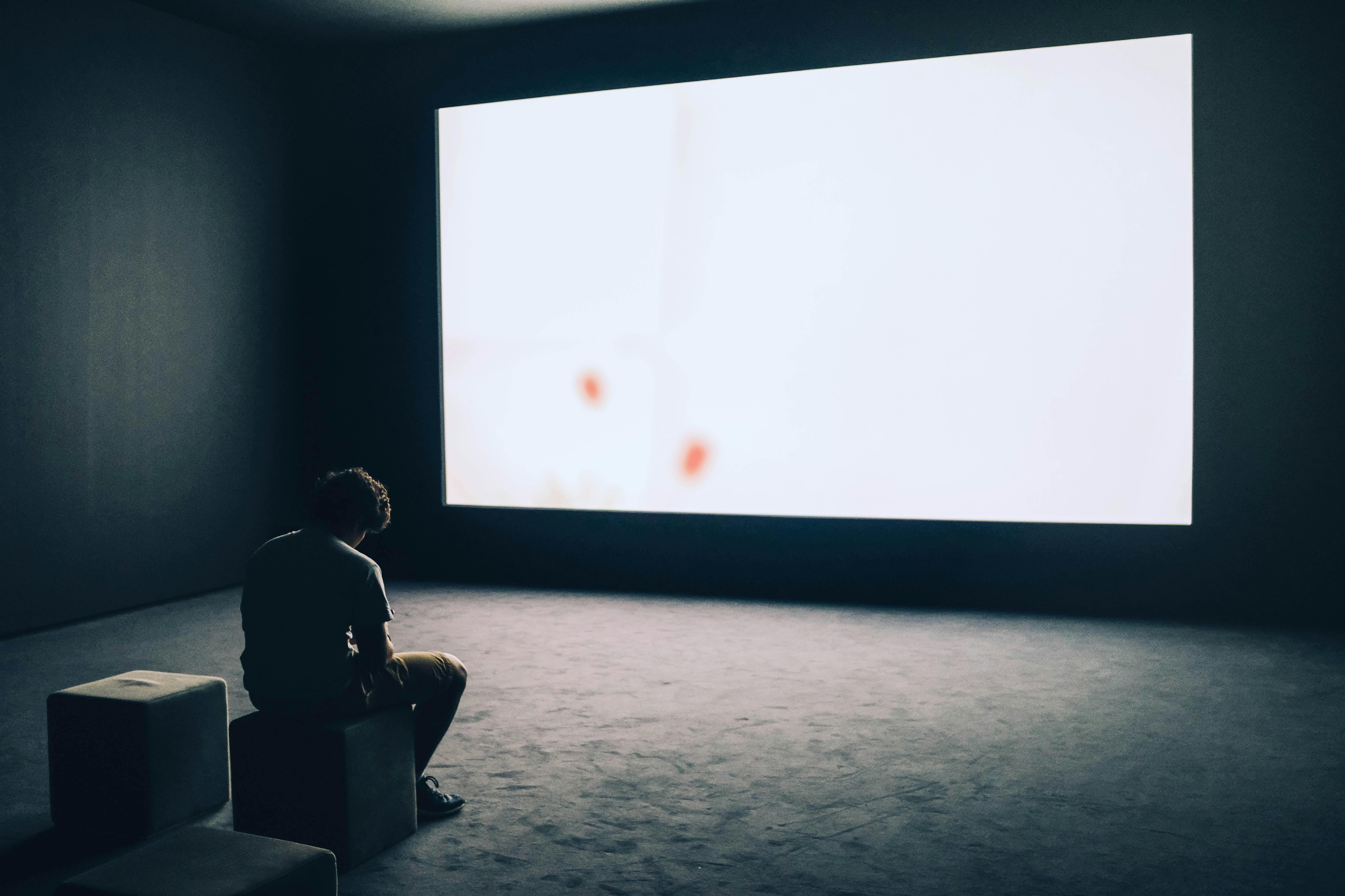 The image shows a young man in a large empty room. He is staring down. In front of him, a massive screen is showing nothing but white. It is the only source of light, casting a cold atmosphere and sharp shadows.