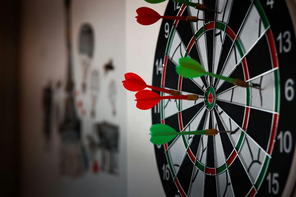 The image shows a dart board. Several darts have pierced the board, with only one in the dead center.