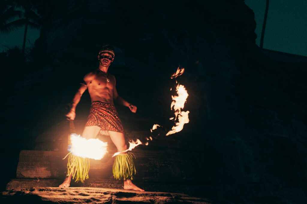 The image depicts a young Samoan man performing the siva afi, a fire dance. He is lit by the fire and ist standing on top of a rockface.