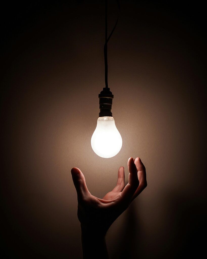 The image shows a close up of a hand reaching for a lit light bulb in an empty room. The light bulb is the only source of light.