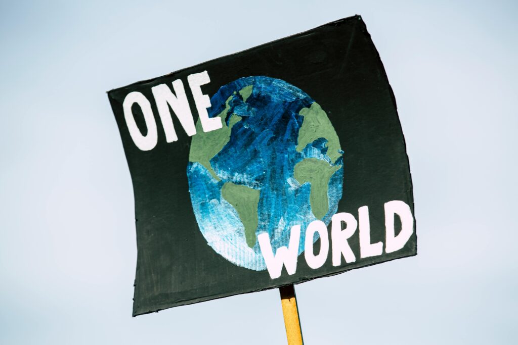 The image depicts a protest sign. The sign features a painting of earth against a black background. The top text says ONE. The bottom text says WORLD. 