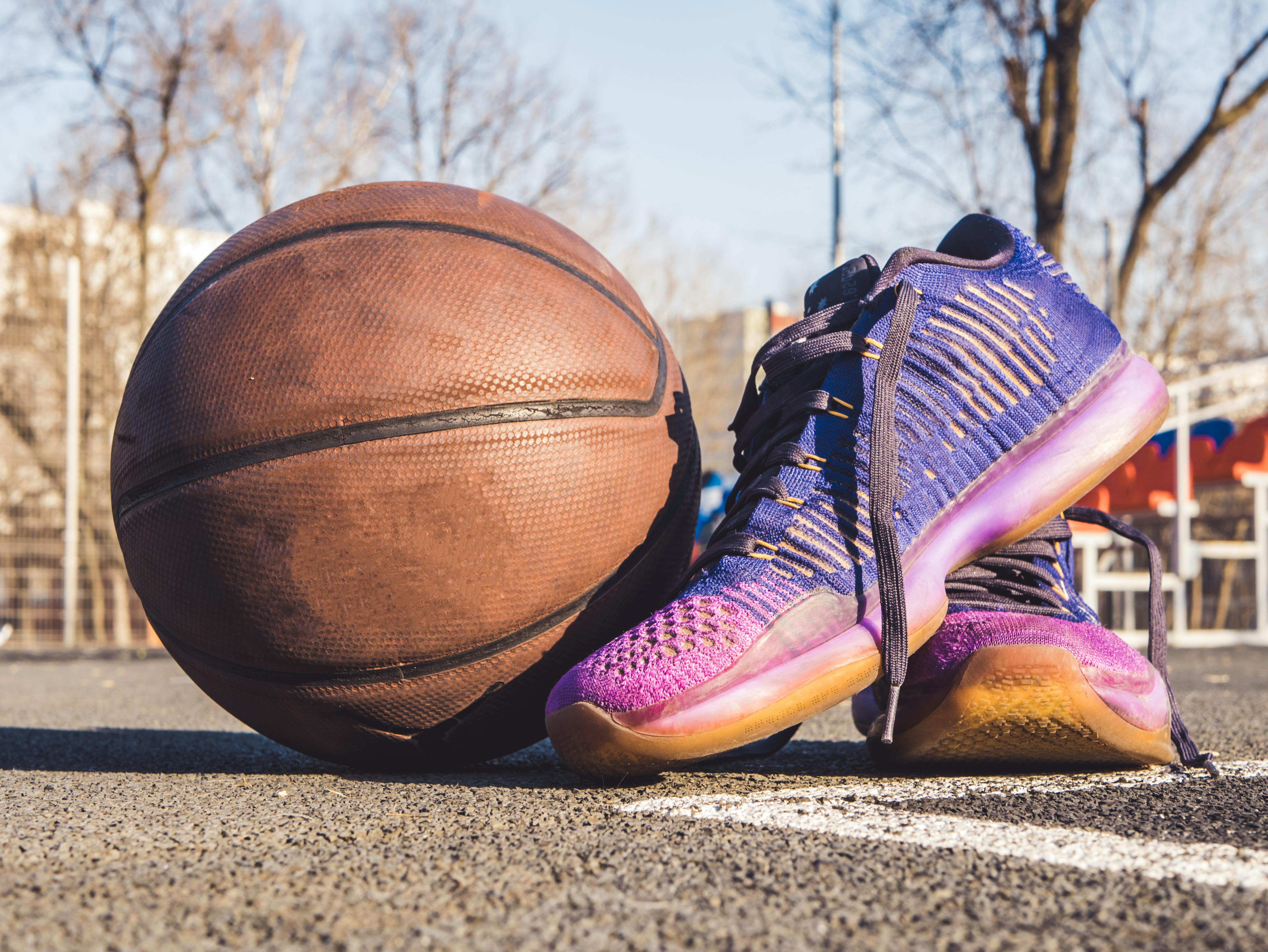 The image shows a dirty basketball next to a clean pair of sports shoes. The shoes are purple, with the soles colored a mix of neon pink and orange. The basketball and shoe are on the asphalt of a basketball court. There are blurred trees and buildings in the background.