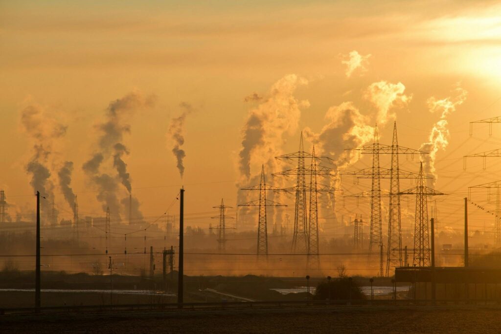 The image shows a city in the background. The stark sunset lighting emphasizing the massive amounts of smoke filling the air. In the foreground is a field, with several electrical towers and wires. The contrast emphasizes the growing threat of climate change