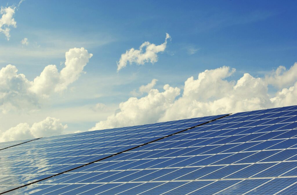 The image shows a large solar panel with cloudy blue skies above it in the background. The rest of the sky is reflecting off the panels in the foreground.