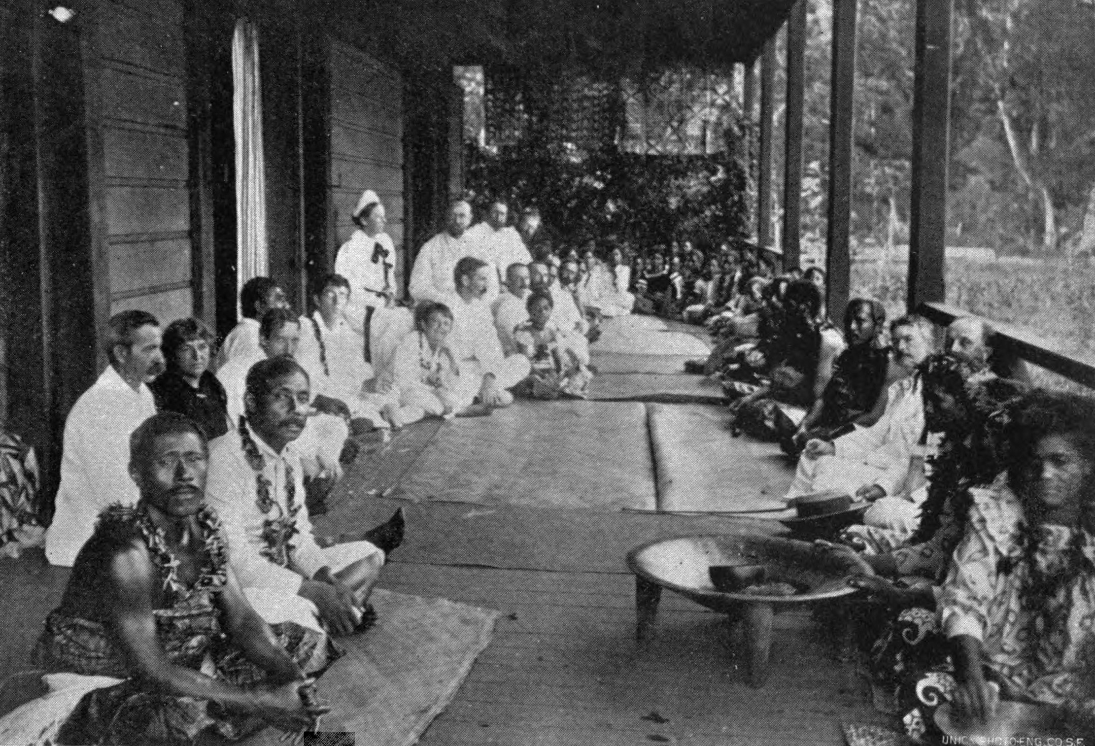 A historical black and white image depicting a gathering of Samoan and European people. Near the center in white, flanked by islanders and Europeans alike, sits Robert Louis Stevenson